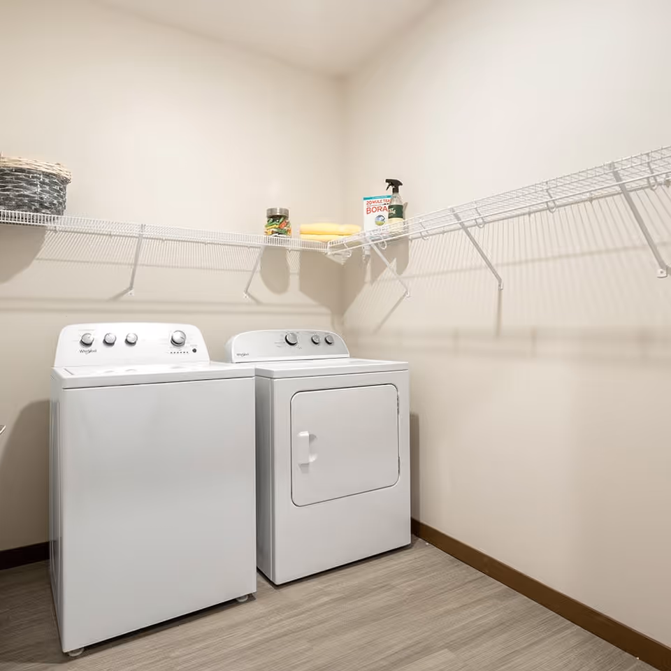 A laundry room with a white washing machine and dryer side by side. Above them is a white wire shelf holding a basket, a jar, folded towels, a box of borax, and a spray bottle. The room has light-colored walls and wood flooring.