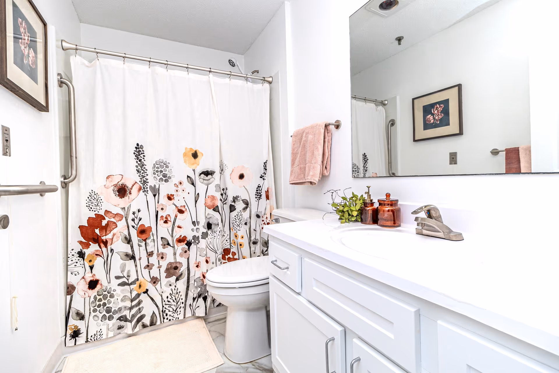 A clean and bright bathroom featuring a white vanity with a sink, a large mirror above it, and a toilet next to a bathtub with a floral-patterned shower curtain. There is a pink towel hanging on a towel rack and a framed floral picture on the wall.