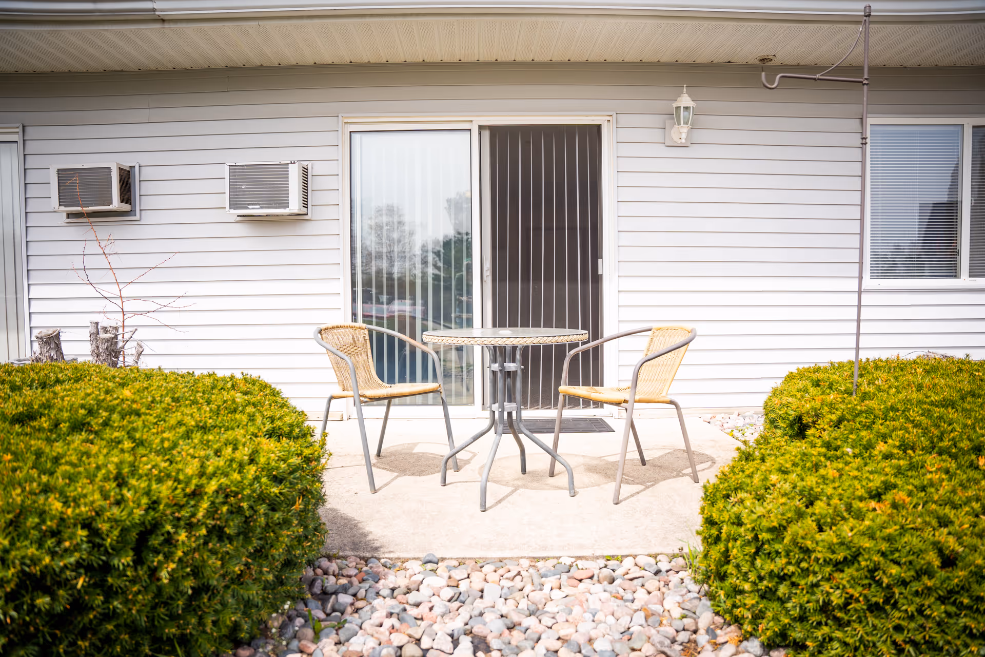 Small patio with two wicker chairs and a round table in front of a sliding glass door, flanked by bushes and wall-mounted air conditioners.