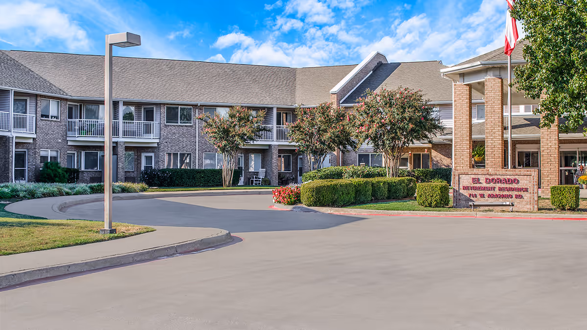 Front driveway and entrance of a two-story senior living building with balconies, landscaping, and a brick sign reading "El Dorado".