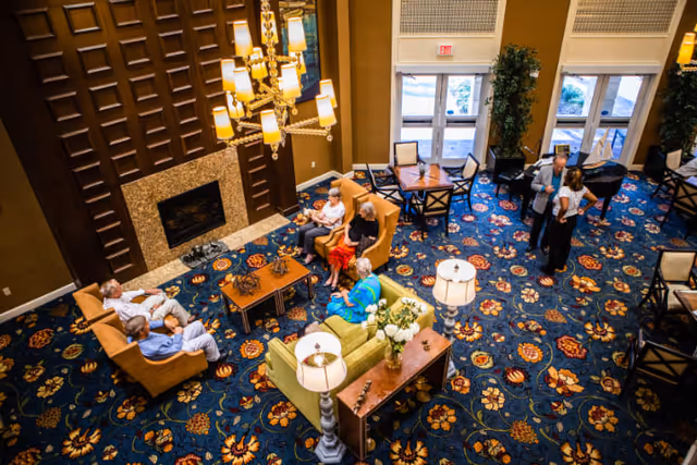 A spacious senior living facility common area with a patterned blue carpet, multiple seating arrangements including sofas and armchairs, a fireplace, and a chandelier. Several elderly people are seated and conversing, while two staff members stand and talk near the entrance doors with large windows.
