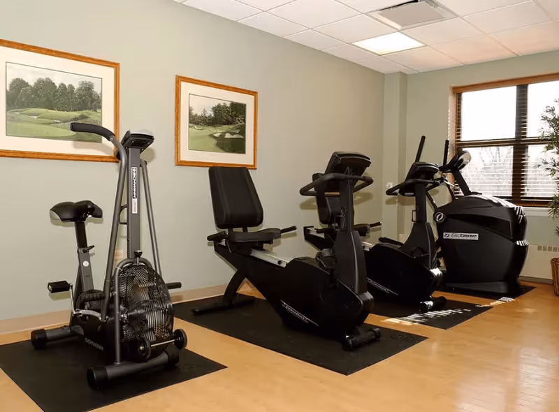 Indoor fitness room with exercise equipment including a stationary bike, two recumbent bikes, and an elliptical machine. The room has light-colored walls with two framed pictures of golf courses, a window with blinds, and a potted plant in the corner.