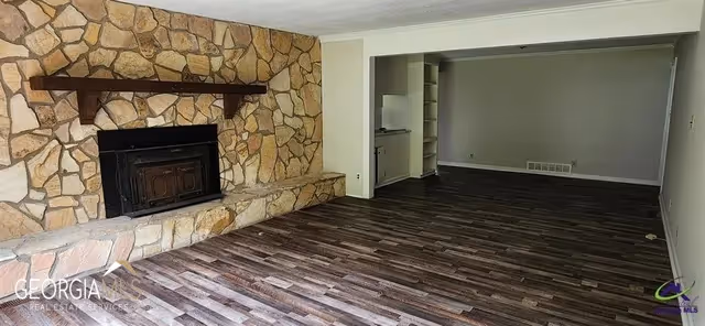 An empty living room with dark wood flooring and a large stone fireplace on the left wall. There is a wooden mantel above the fireplace. The room extends into an adjacent area with built-in shelves and a small kitchenette visible in the background. The walls are painted light gray.