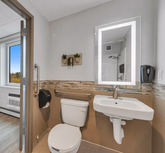 A clean and modern bathroom with a white toilet and a wall-mounted sink. There is a large illuminated mirror above the sink, a grab bar next to the toilet, and a small decorative wall piece with plants. The bathroom has beige and stone tile accents, and an open door reveals a room with a window and wooden flooring.