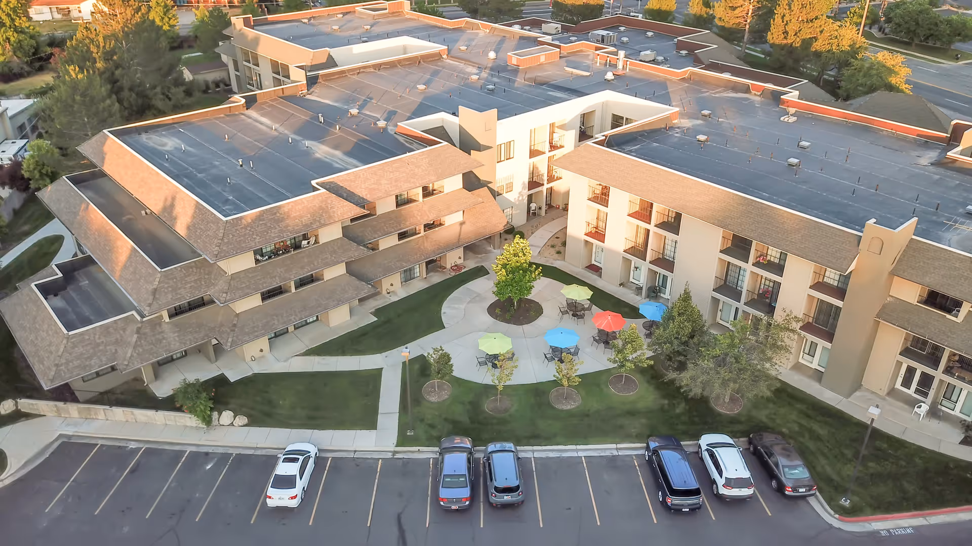 Aerial view of Cottonwood Creek senior living facility showing a multi-story building with balconies, a central courtyard with colorful patio umbrellas and seating, surrounded by green lawns and trees, and a parking lot with several cars parked.