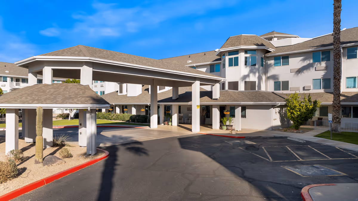Exterior view of Vista del Rio - A Provincial Senior Living Community showing the entrance with a covered drop-off area, multiple windows, and a clear blue sky. There are desert plants and a paved driveway with parking spaces in front of the building.