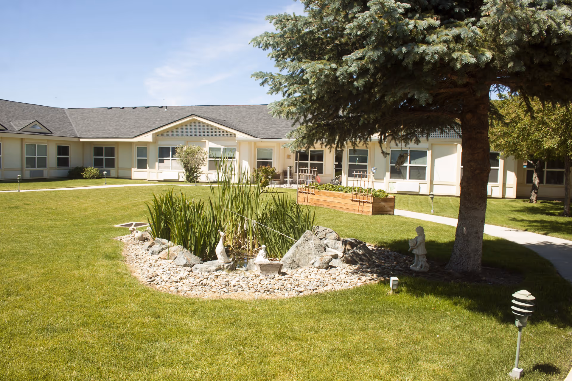 Outdoor courtyard area of Settler's Park Senior Living featuring a small pond with rocks and plants, a large tree providing shade, a garden bed with trellises, and a surrounding walkway with a single-story building in the background under a clear blue sky.