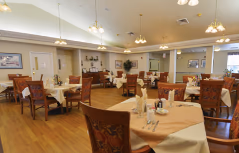 A spacious dining room with multiple tables covered in beige tablecloths and set with napkins, glasses, and utensils. The room has wooden floors, decorative chairs with patterned upholstery, and soft overhead lighting. There are framed pictures on the walls and some plants for decoration.