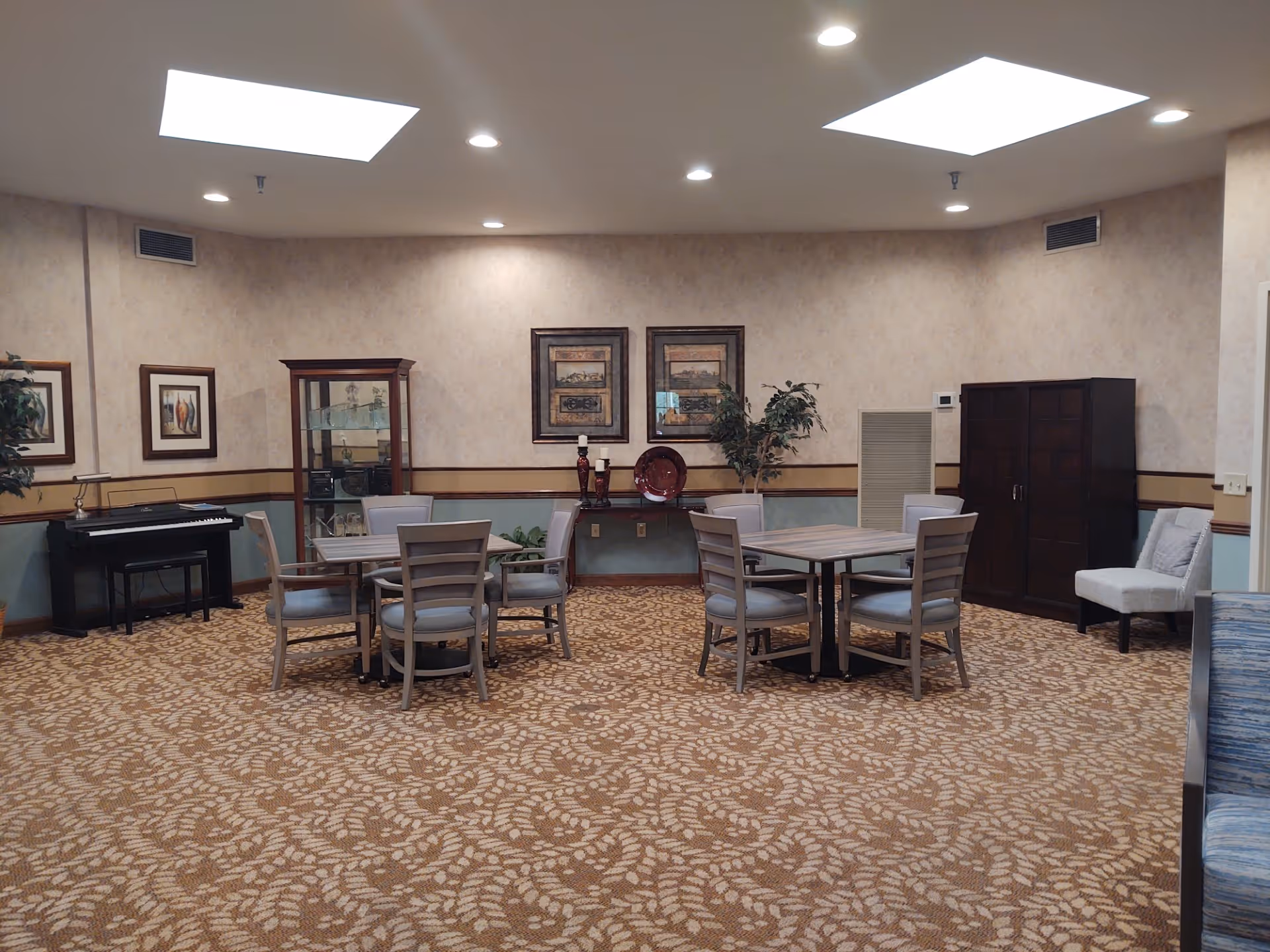 A senior living common room with round tables and chairs, a piano, cabinets and framed artwork under skylights.