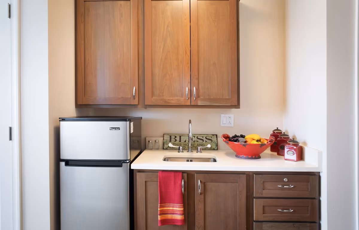 A small kitchenette area with wooden cabinets, a stainless steel mini refrigerator, a sink with a faucet, a red dish towel hanging from a cabinet handle, a decorative sign that reads 'BLESS THIS HOME AND ALL WHO ENTER', a red bowl filled with fruit, and three red canisters on the countertop.