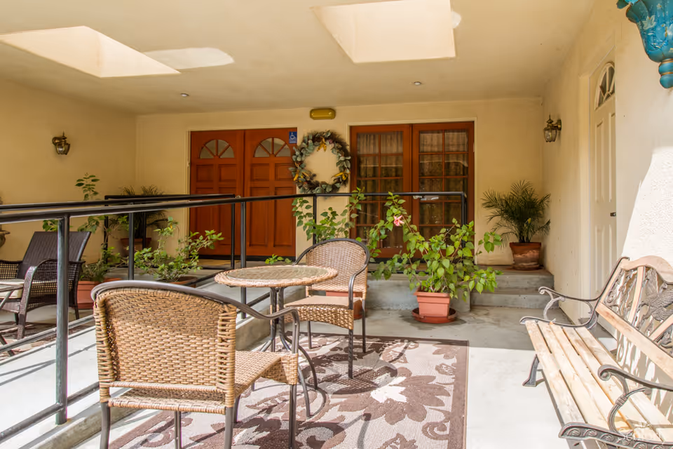 Covered outdoor patio area with wicker chairs and a round table on a floral patterned rug. There are potted plants and a wooden bench with metal armrests. Two wooden doors and a glass door are visible in the background, along with a decorative wreath hanging on the wall.