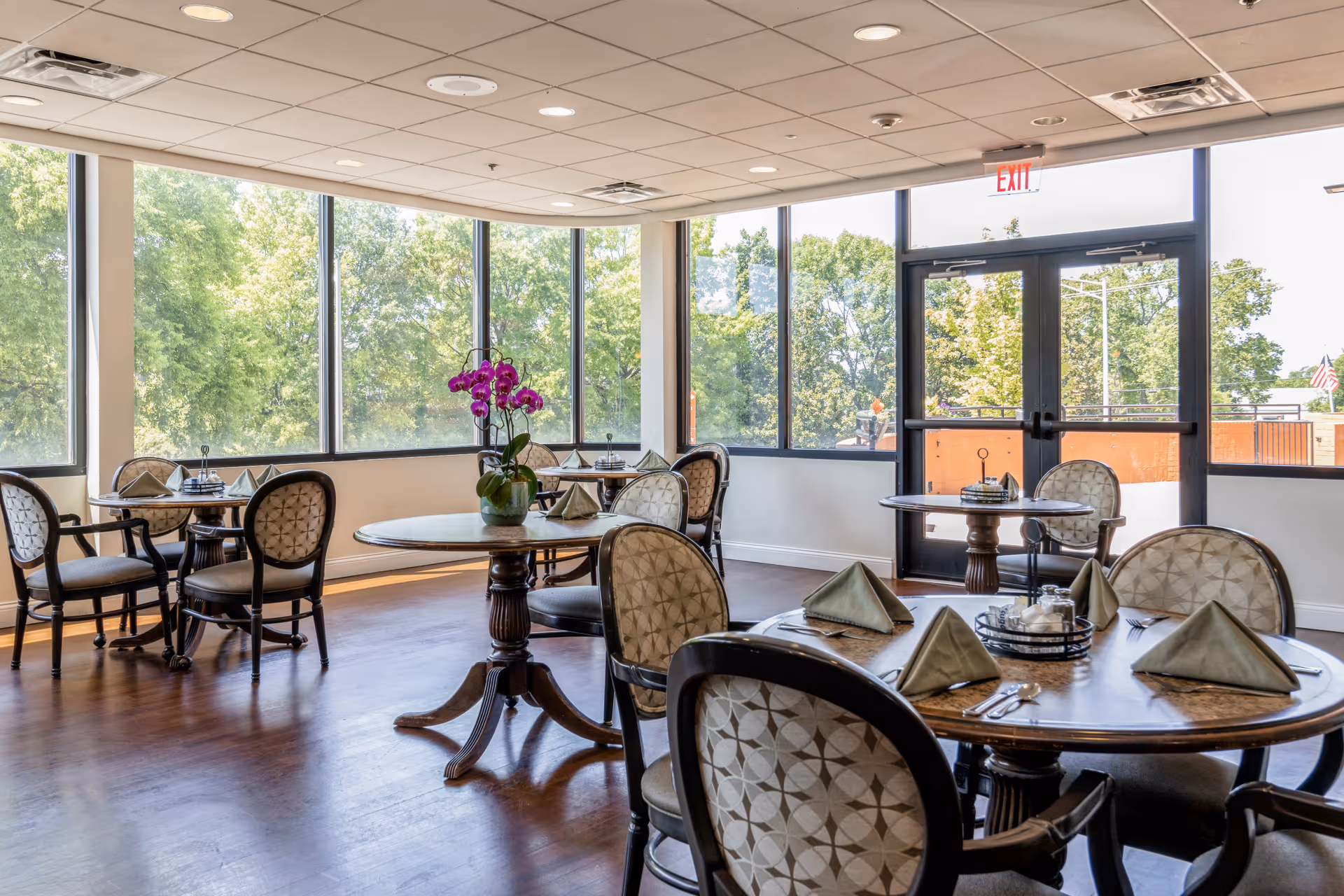 A bright dining room with round wooden tables set with folded napkins, silverware, and condiments. The room has large windows letting in natural light and offering a view of green trees outside. There is a purple orchid centerpiece on one of the tables, and an exit door with glass panels is visible.