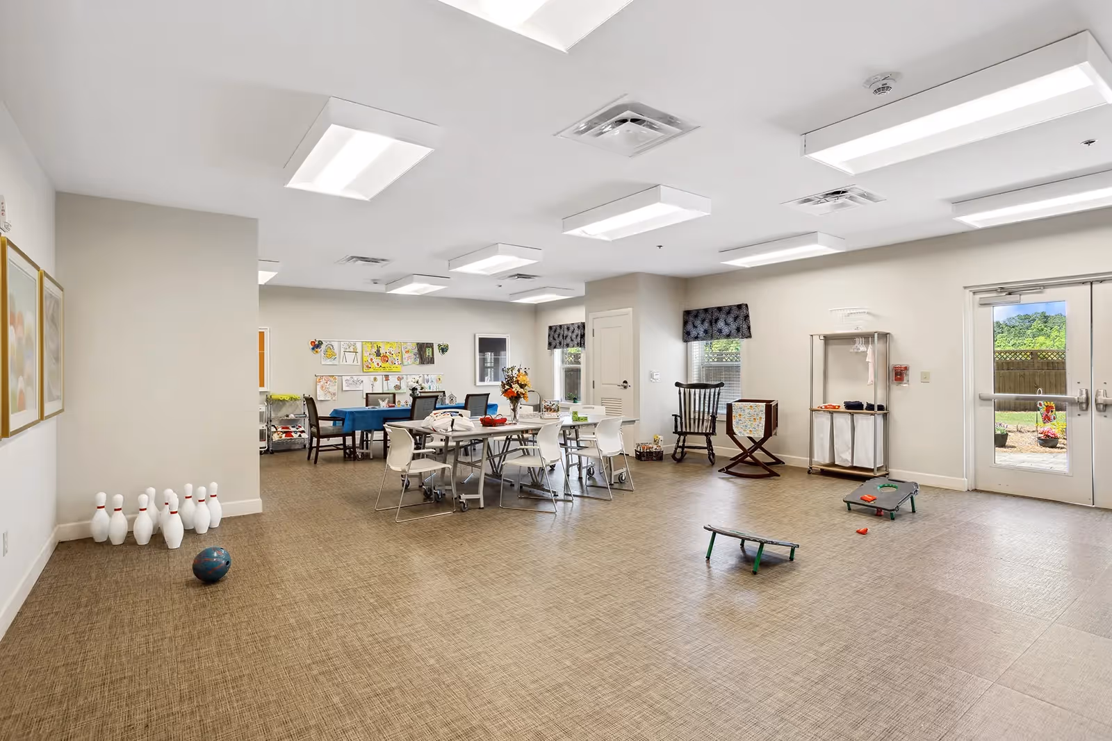 A spacious activity room in a senior living facility with tables and chairs arranged for group activities. There are bowling pins and a bowling ball set up on the floor, a cornhole game, and a rocking chair near the windows. The room has bright overhead lighting, light-colored walls, and a door leading outside to a fenced area with greenery.