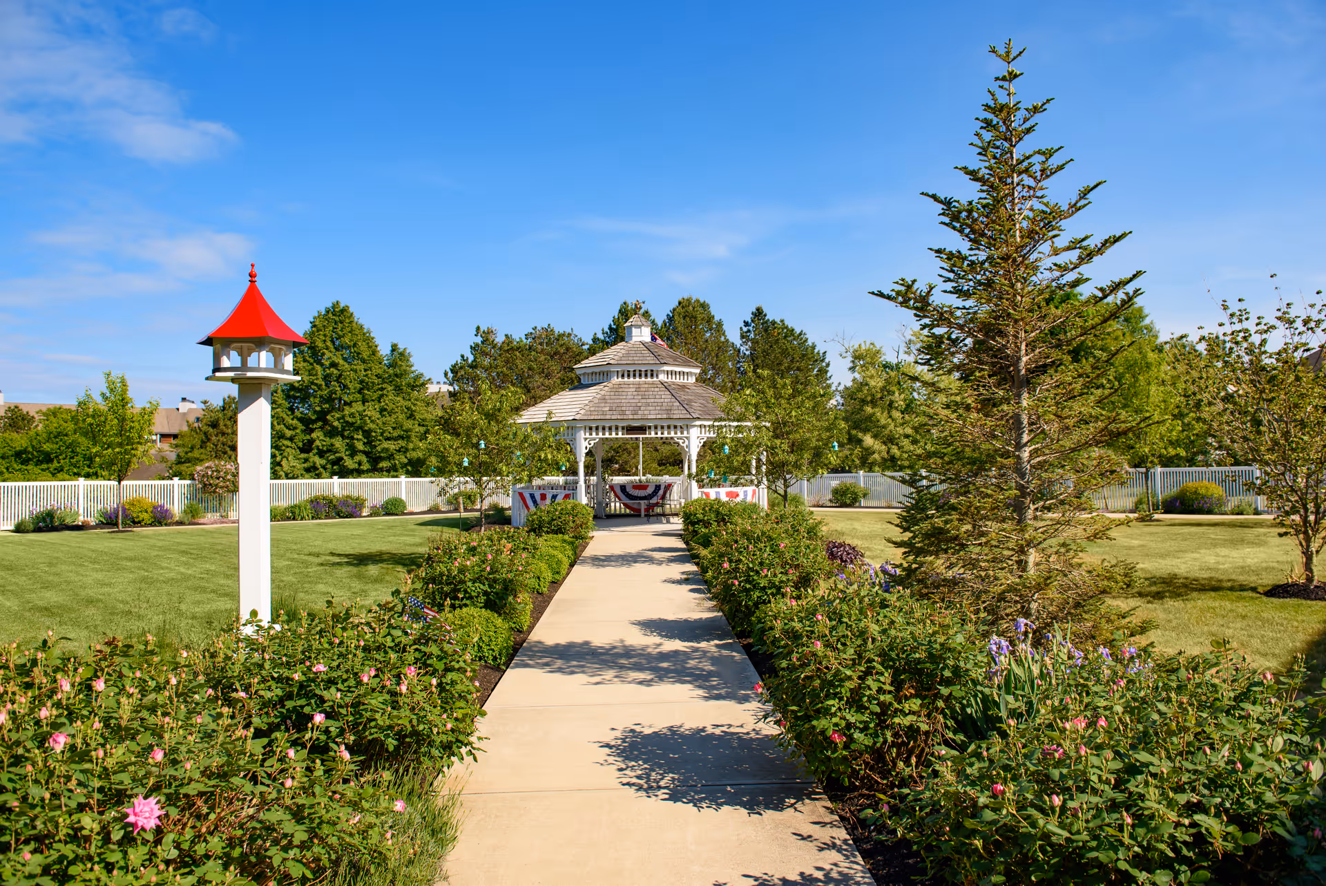 A paved walkway through a landscaped lawn leads to a white gazebo surrounded by trees and flowerbeds under a blue sky.