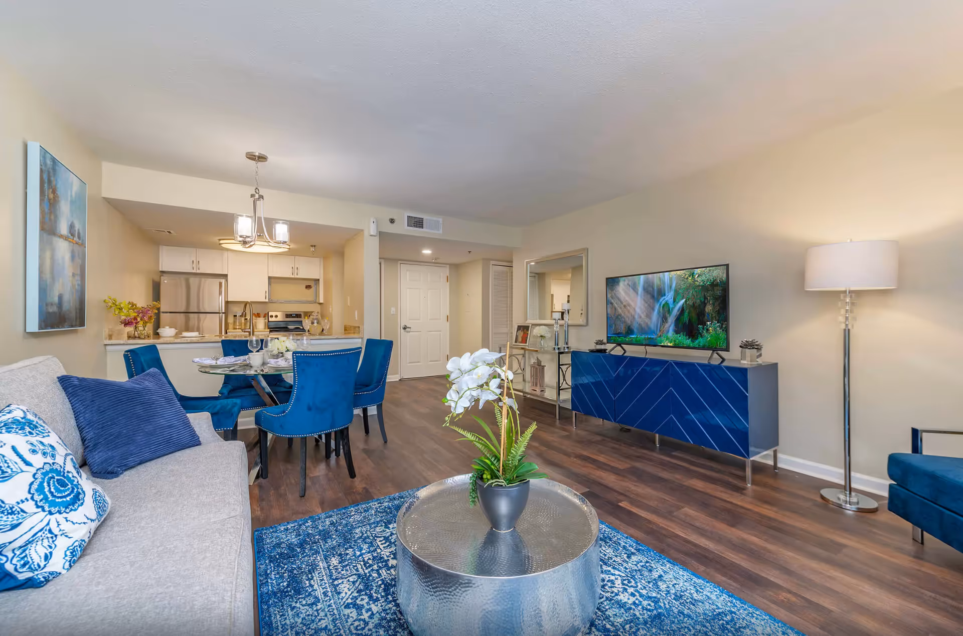 A modern living room and dining area in a senior living facility featuring a gray sofa with blue and white pillows, a round metallic coffee table with a potted white orchid, a blue patterned rug, a dining table with blue upholstered chairs set for four, a blue TV console with a flat-screen TV displaying a waterfall scene, a floor lamp, and a kitchen with stainless steel appliances in the background.