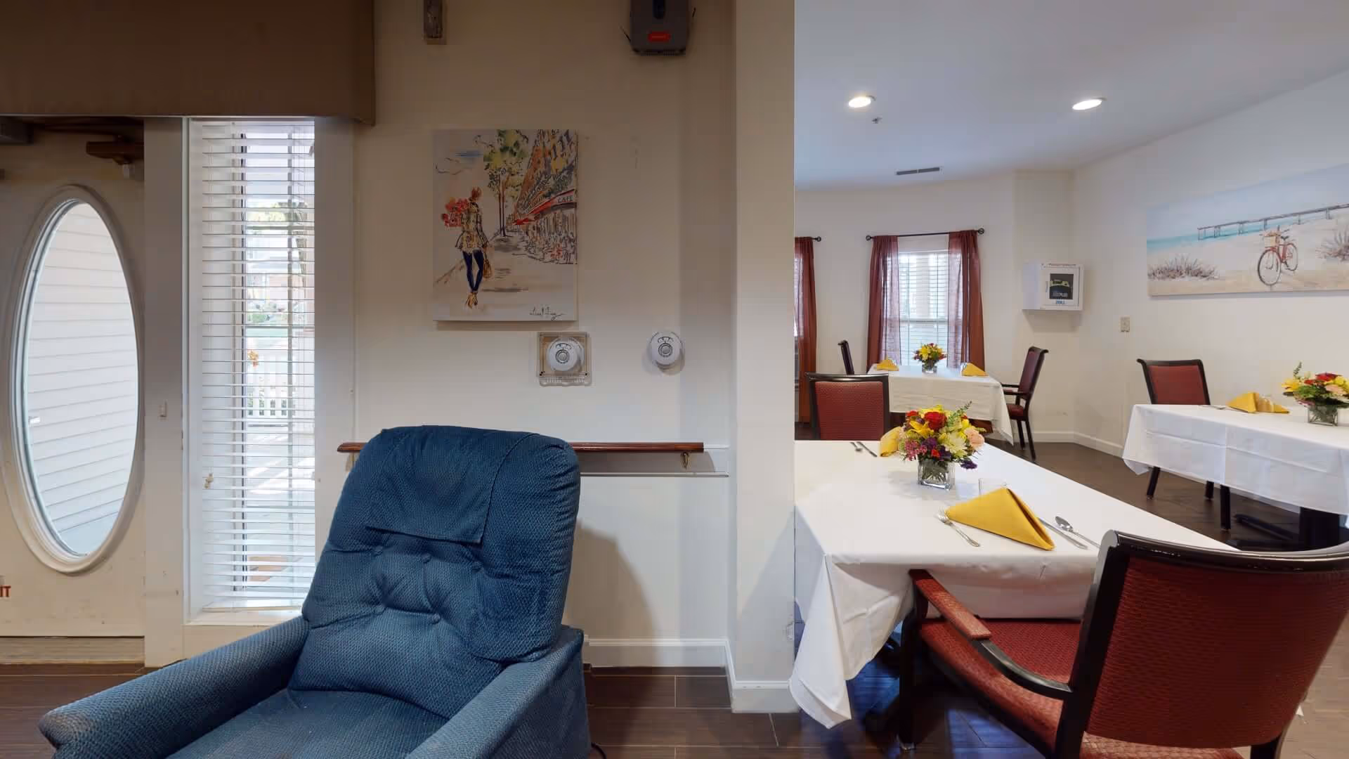 A common area with a blue recliner in the foreground and dining tables set with white tablecloths and yellow napkins.