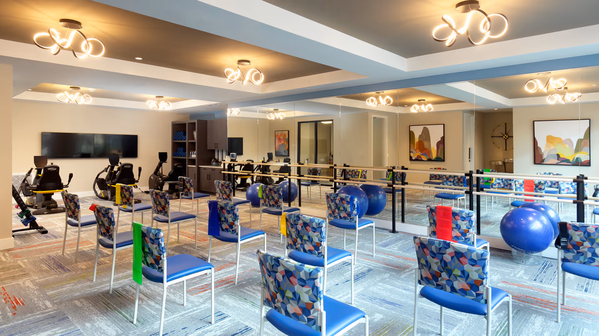 A bright exercise room with multiple chairs arranged in rows, each with colorful resistance bands hanging on the back. The room features large mirrors on one wall, exercise balls, and stationary exercise bikes. Modern ceiling lights illuminate the space, and there are abstract paintings on the walls.