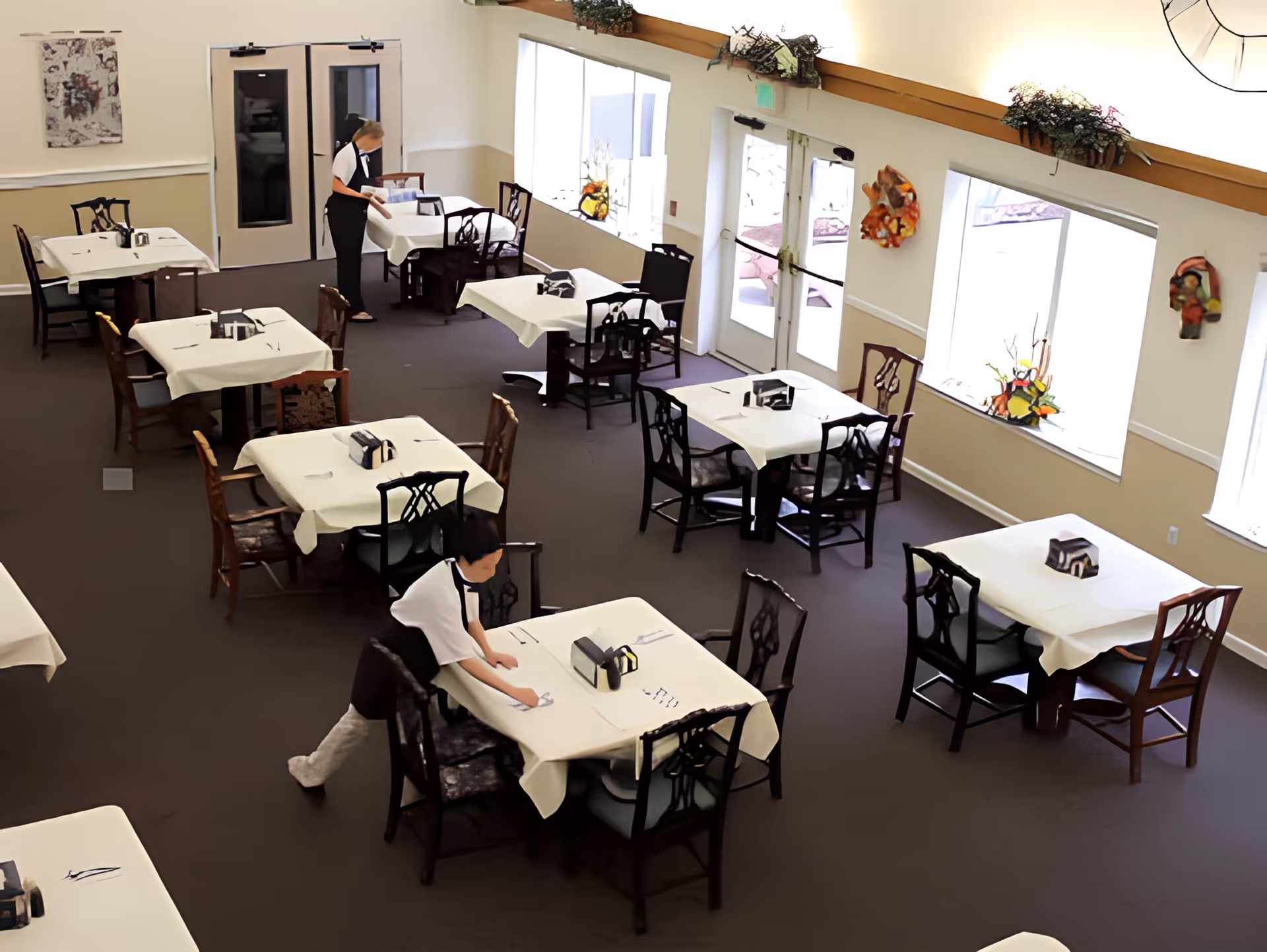 A dining room in an assisted living community with several tables covered in white tablecloths. Two staff members are setting tables with utensils and napkins. Large windows and glass doors let in natural light, and decorative wall hangings are visible.