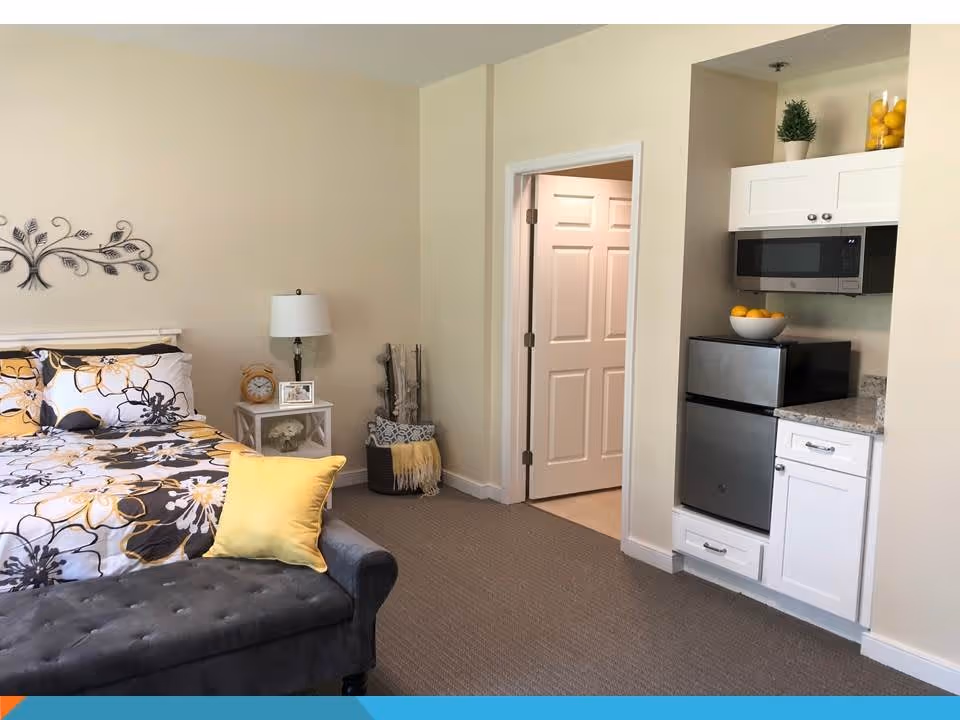 A cozy bedroom featuring a bed with floral patterned bedding in shades of yellow, black, and white, a gray tufted bench at the foot of the bed, a small white nightstand with a lamp, clock, and picture frame, and a small kitchenette area with a microwave, mini refrigerator, and white cabinets. The room has beige walls and carpeted flooring.