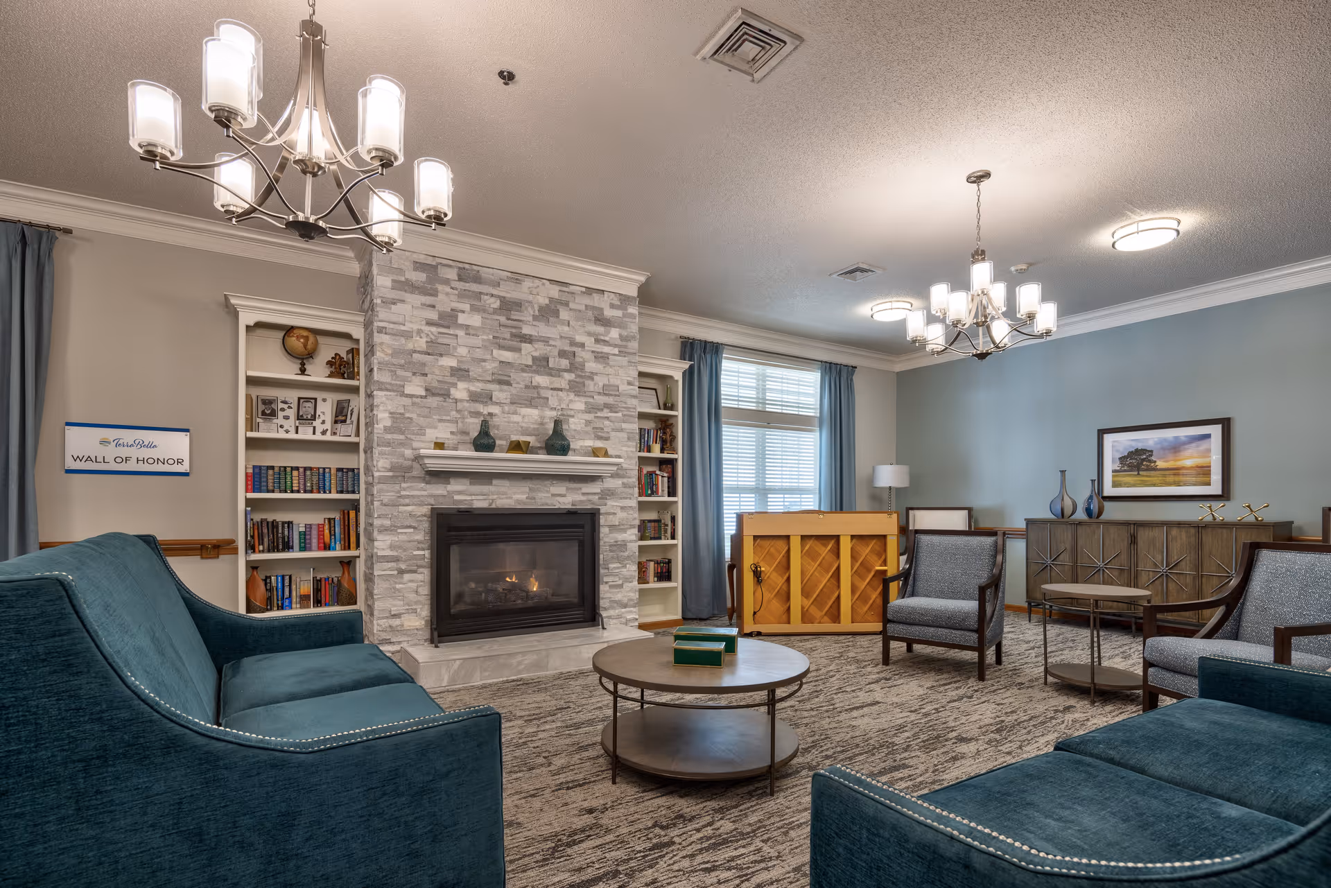 A cozy living room with teal upholstered sofas and armchairs arranged around a round coffee table. The room features a stone fireplace with a mantle decorated with vases, built-in bookshelves filled with books and decor, a piano near a window with blue curtains, and a sideboard with decorative items and a framed landscape picture on the wall. Two modern chandeliers hang from the ceiling providing warm lighting.