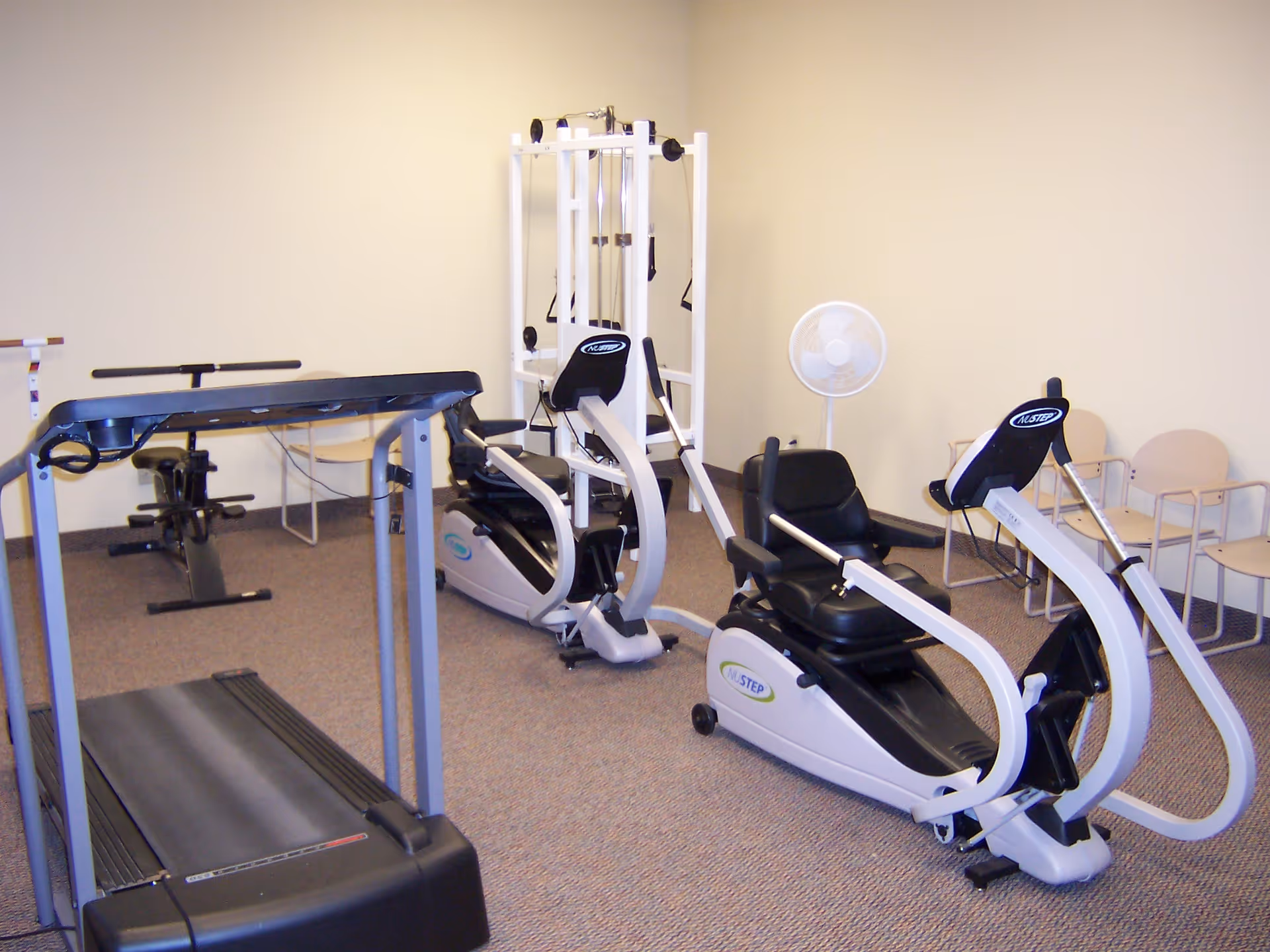 A small exercise room with a treadmill, two recumbent exercise bikes, a multi-function weight machine, a standing fan, and several beige chairs lined up against the wall.