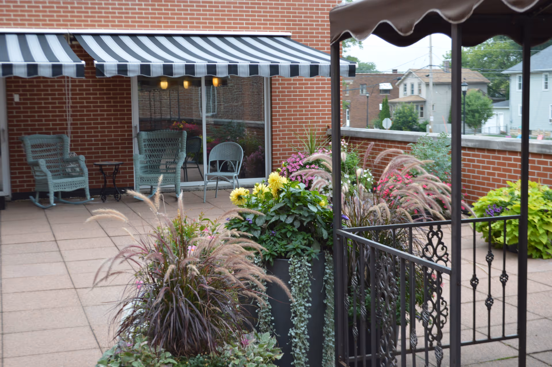 Outdoor patio area with potted plants and flowers, a black metal gazebo structure, brick walls, and a building with striped awnings and green wicker chairs.