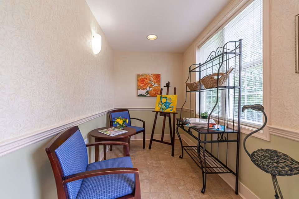 A small sitting area with two blue cushioned wooden chairs and a round wooden table between them holding a magazine and a small vase with yellow flowers. On the right side, there is a black metal shelving unit with baskets and books near a window with blinds. An easel with a colorful painting and a floral painting on the wall are visible in the background. The walls have a light patterned wallpaper with a chair rail and the floor is tiled.