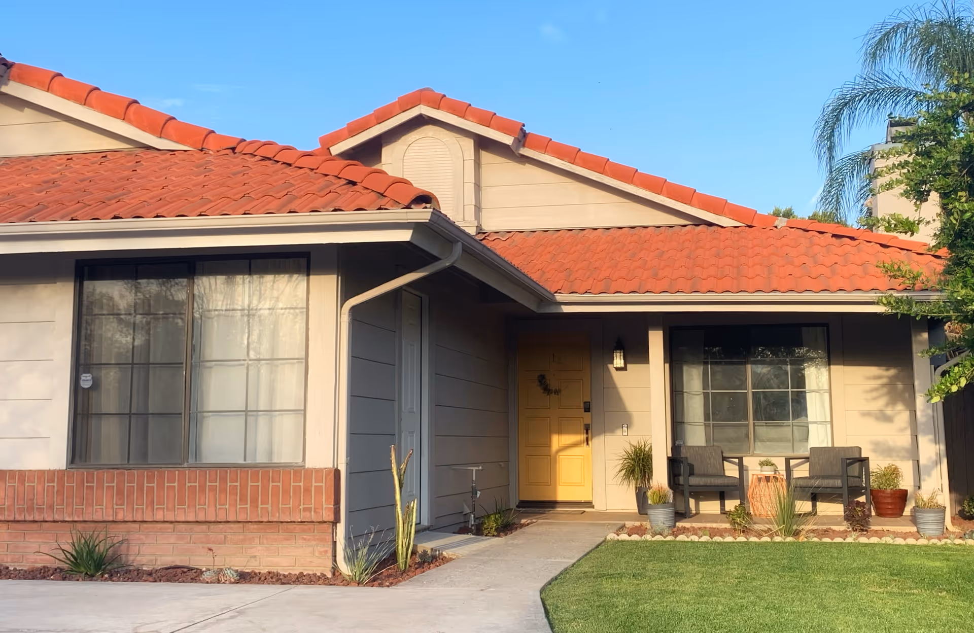 Front exterior view of a single-story house with a red tile roof, beige walls, a yellow front door, two large windows with curtains, a small porch with two chairs and a table, potted plants, and a well-maintained lawn under a clear blue sky.