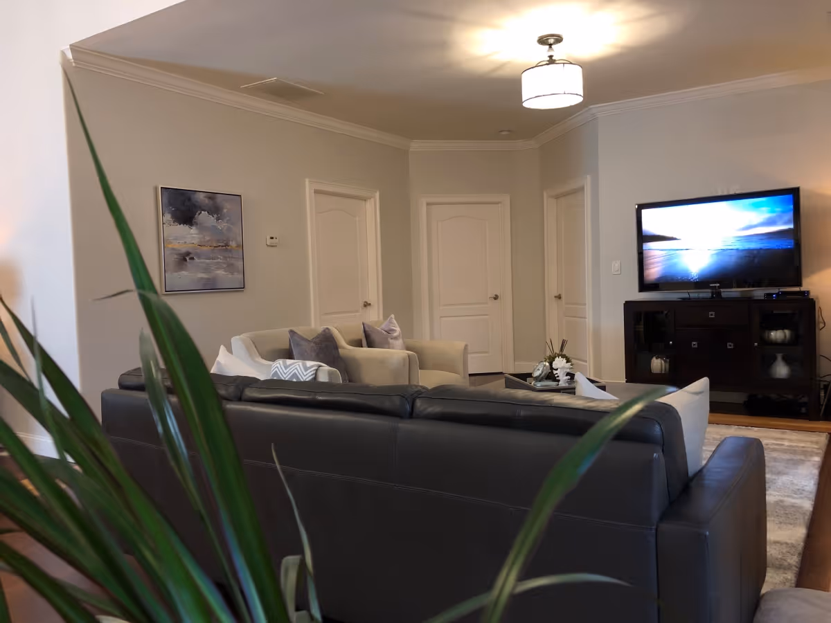 A cozy living room with a dark leather sofa and light-colored armchairs arranged around a coffee table. A flat-screen TV is mounted on a dark wooden cabinet against the wall, displaying a scenic ocean view. There is a modern ceiling light fixture, a framed abstract painting on the wall, and some green plant leaves partially visible in the foreground.