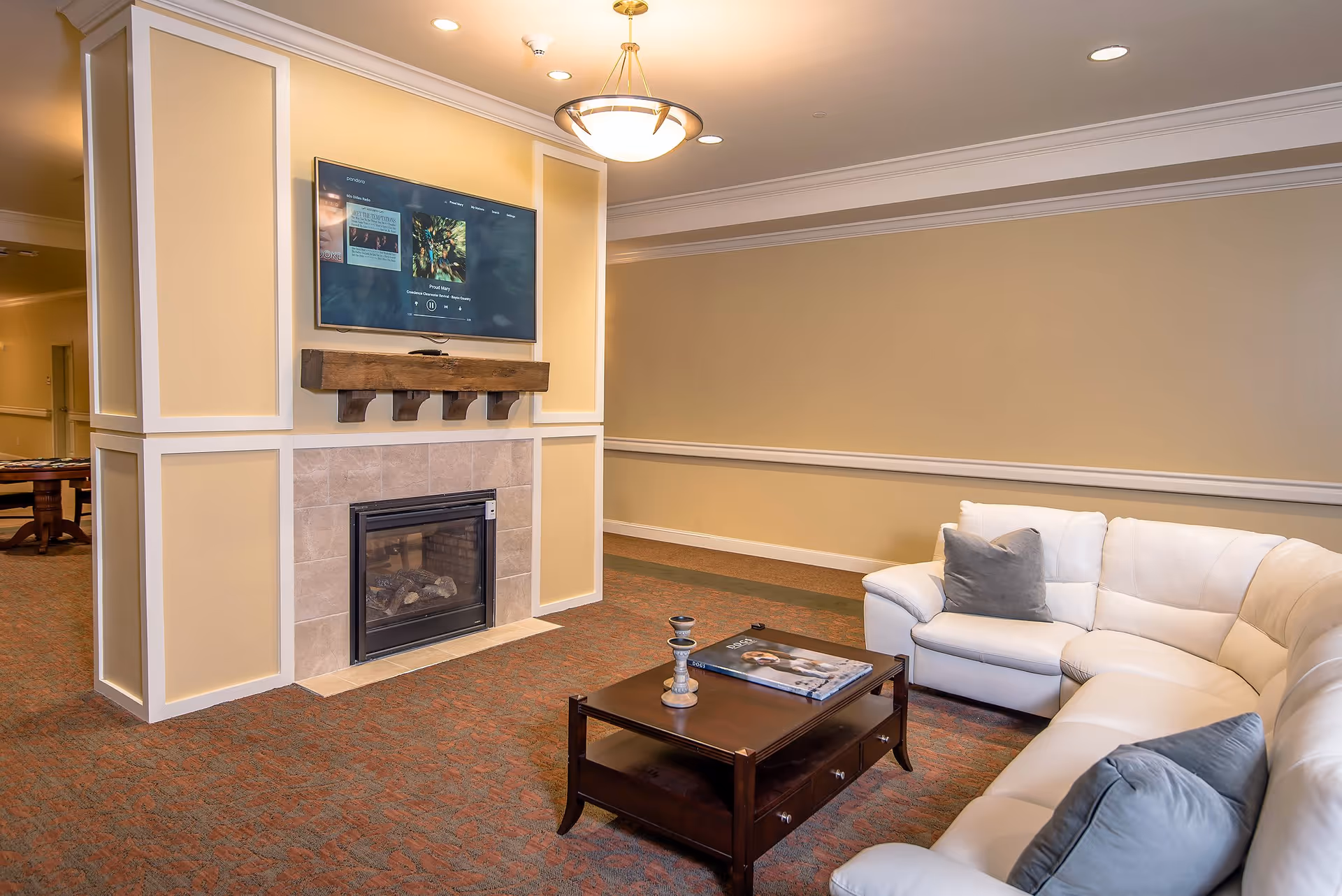 A cozy living room area with a white sectional sofa adorned with gray pillows, a dark wooden coffee table with decorative items and a book, a fireplace with a wooden mantel, and a flat-screen TV mounted above the fireplace. The walls are painted beige with white trim, and the carpet has a patterned design.