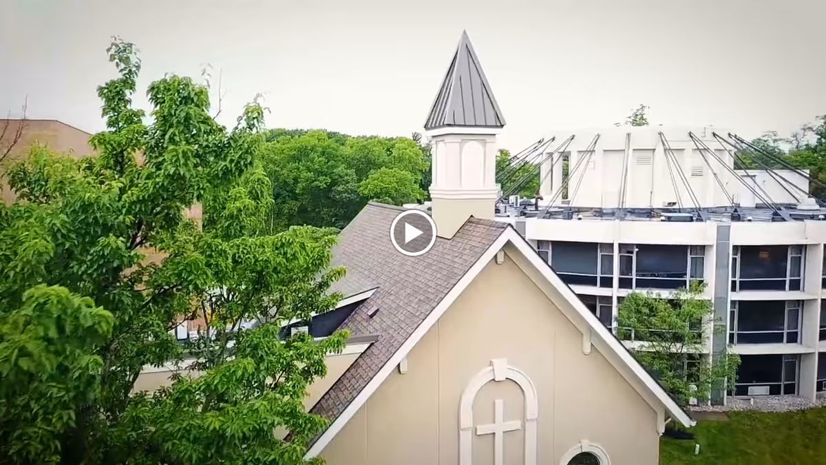 Exterior view of a building with a steep roof and a small tower with a pointed roof, surrounded by green trees and another modern building in the background.