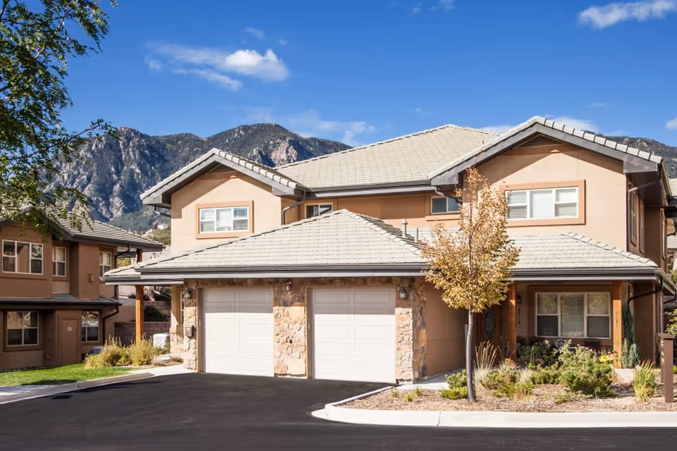 Exterior view of a two-story residential building with beige walls, a tiled roof, and two white garage doors. There is a small landscaped area with a tree and shrubs in front of the building. Mountains and a blue sky with a few clouds are visible in the background.