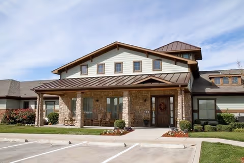 Front exterior view of a single-story assisted living facility with a stone and siding facade, a covered entrance with wooden rocking chairs, and a well-maintained lawn and flower beds under a partly cloudy sky.