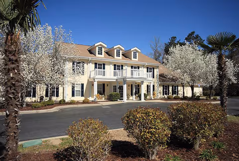 Exterior view of a two-story residential building with a beige facade, white trim, and a covered entrance. The building is surrounded by landscaped bushes, palm trees, and flowering trees under a clear blue sky.