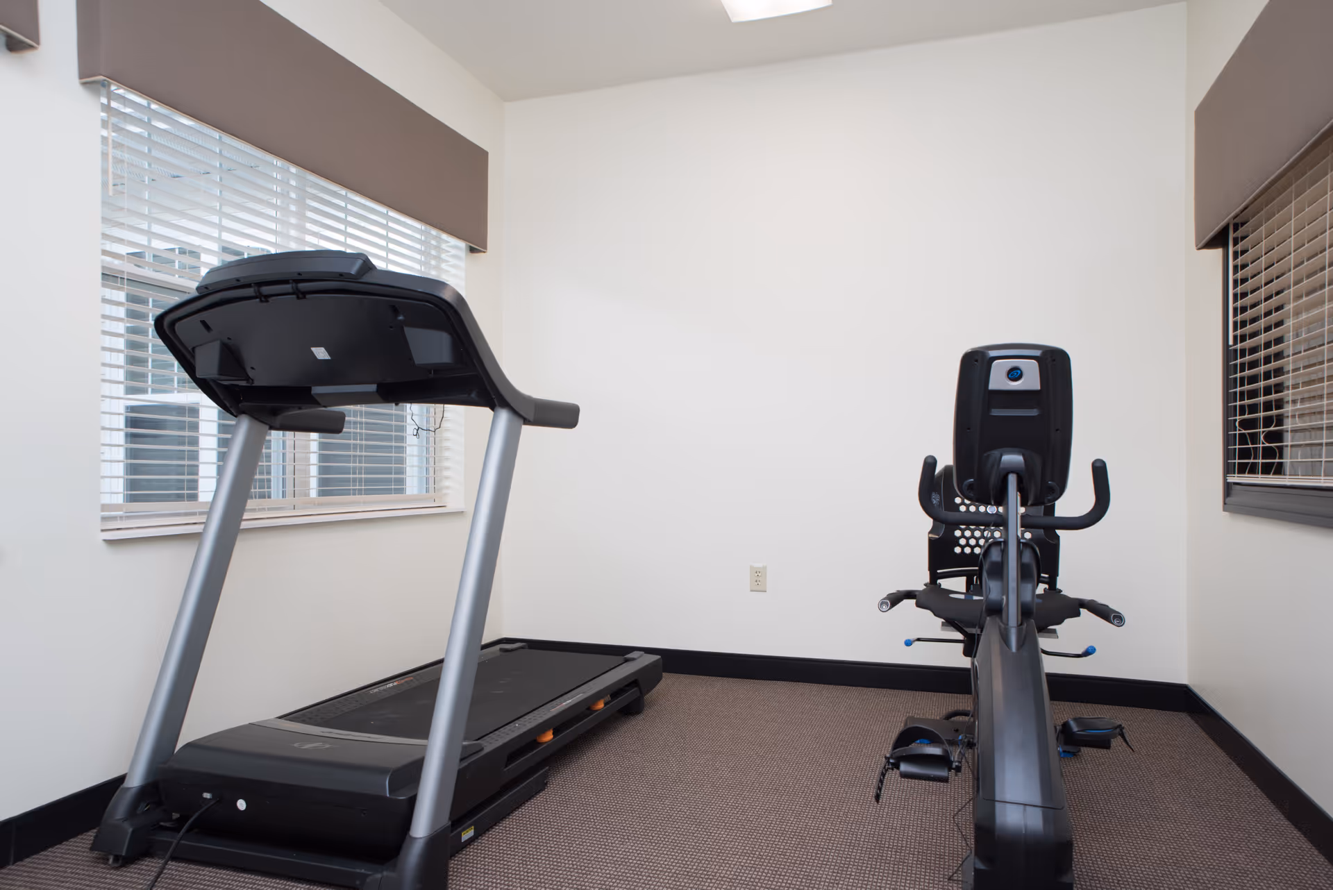 A small exercise room with a treadmill on the left and a recumbent stationary bike on the right. The room has beige walls, two windows with blinds and brown valances, and a carpeted floor.