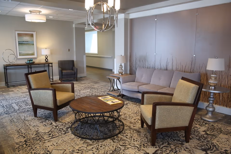A cozy living room area with a beige sofa, two armchairs with wooden frames and patterned upholstery, a round wooden coffee table with a metal base, and two side tables each with a lamp. The room has a patterned carpet, a modern chandelier, a framed abstract painting on the wall, and a console table with decorative items.