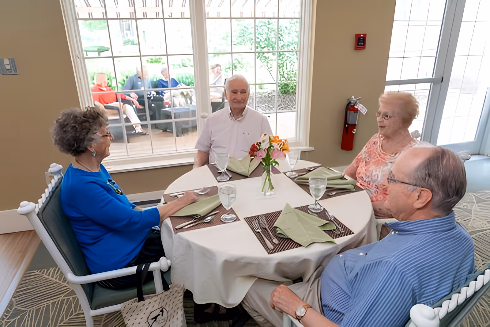 Four elderly people sitting around a round dining table set with placemats, folded green napkins, and glasses of water. There is a small vase with flowers in the center of the table. Large windows behind them show more elderly people sitting outside on a patio.