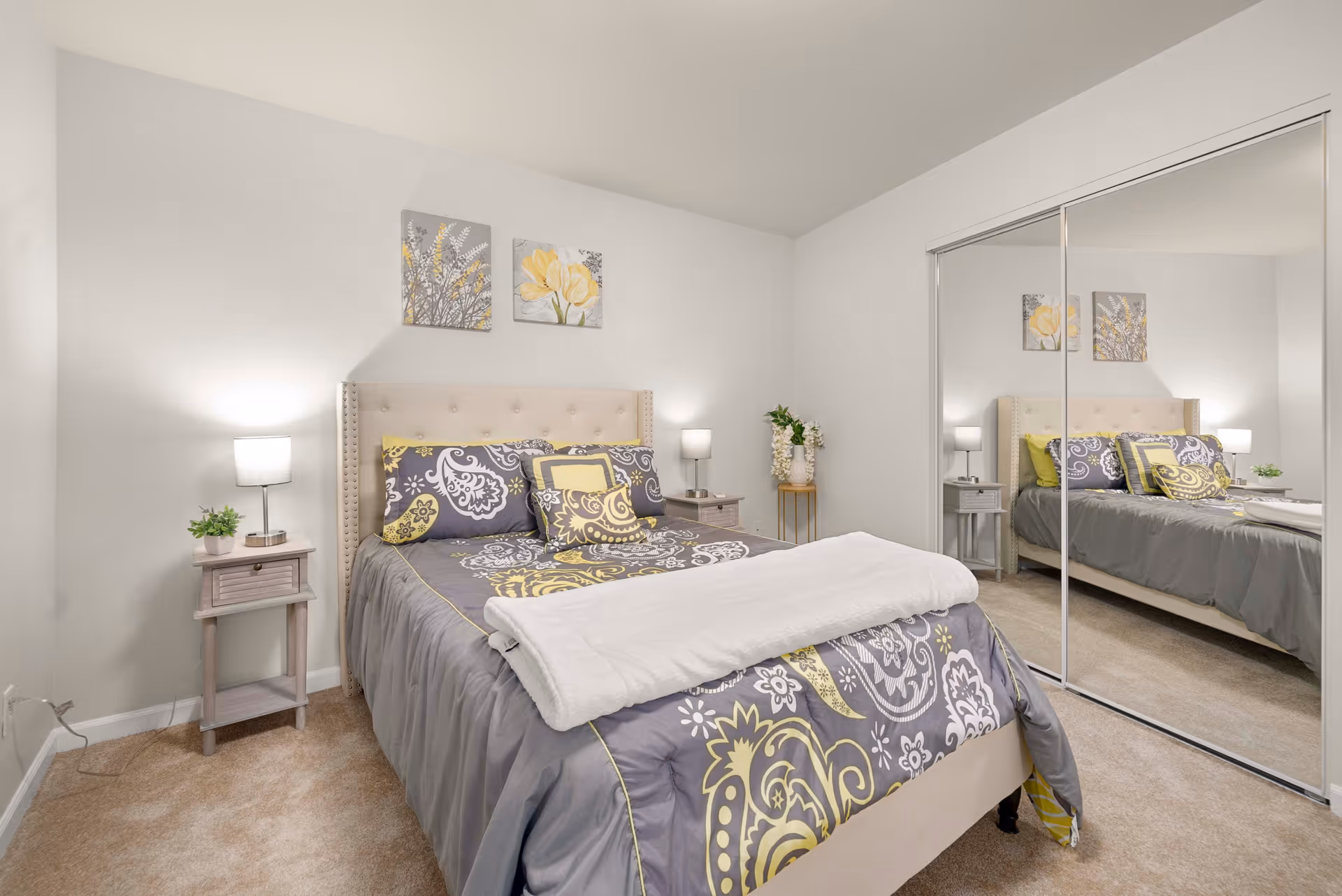A neatly made bed with a gray and yellow patterned comforter and pillows in a bedroom. The bed has a beige tufted headboard. There are two small nightstands on either side of the bed, each with a lamp and a small plant. Above the bed, two floral paintings hang on the white wall. A large mirrored sliding closet door is on the right side of the room, reflecting the bed and nightstands. The room has beige carpet flooring and white walls.