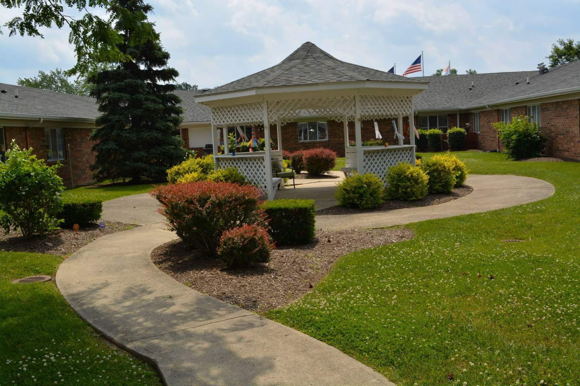 Well-maintained courtyard featuring a white lattice gazebo, winding concrete path, shrubs, and single-story brick buildings in the background.
