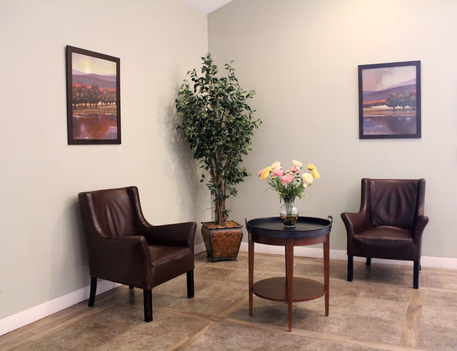 Two brown leather chairs flank a small round table topped with a vase of flowers, set against a pale wall with framed artwork and a potted plant.