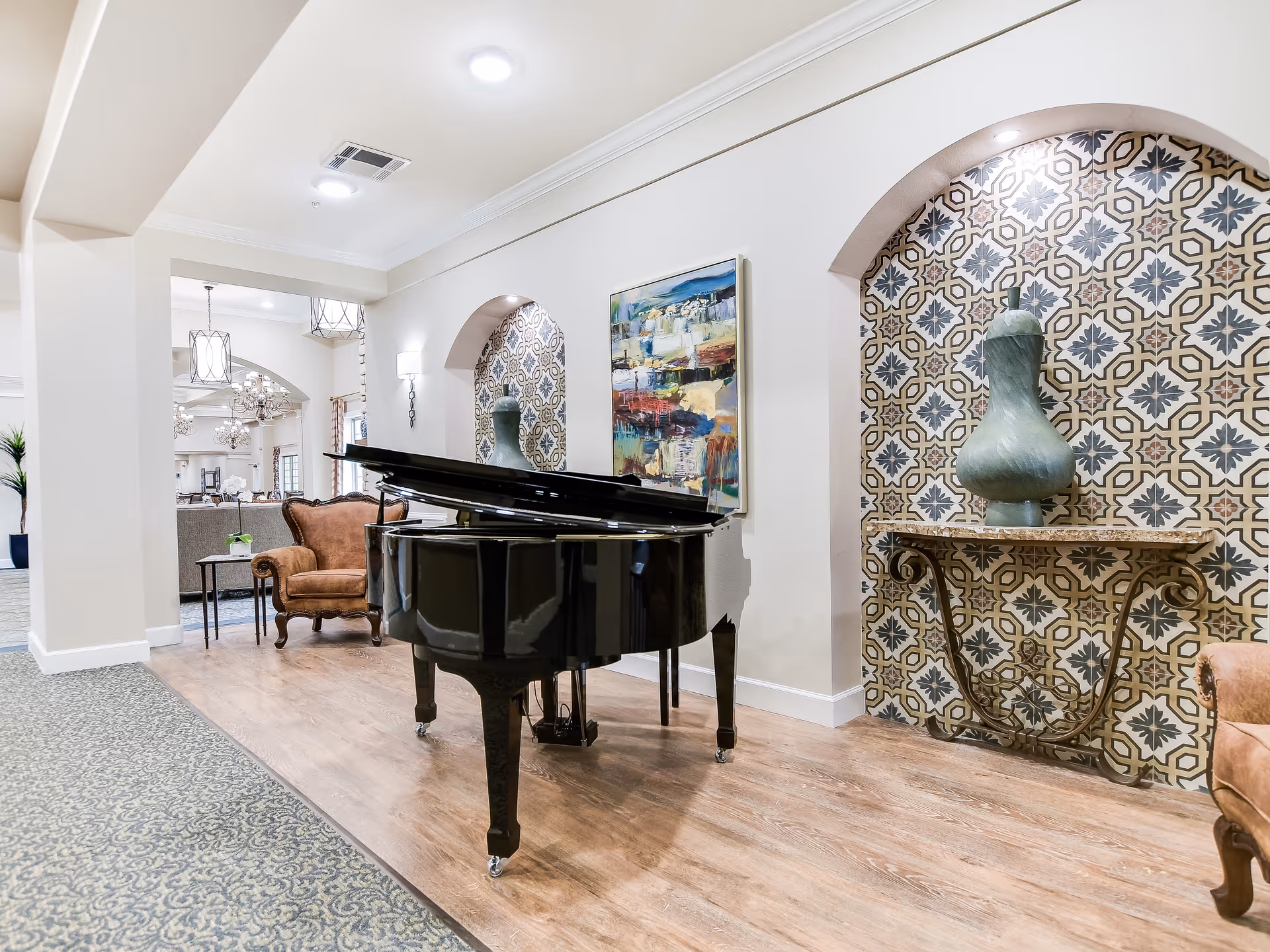 A black grand piano sits in a bright senior living common room flanked by upholstered chairs and patterned decorative alcoves.