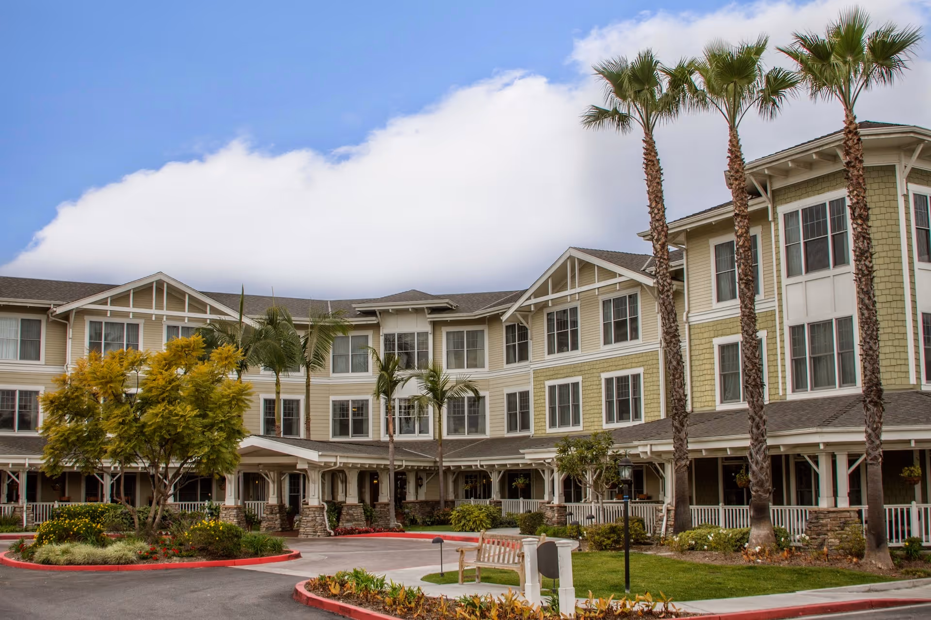 Exterior view of a multi-story senior living facility with beige and white siding, numerous windows, palm trees, and landscaped greenery under a partly cloudy sky.