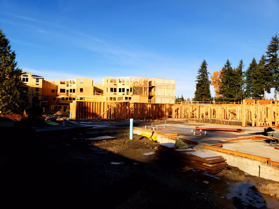 Construction site of a multi-story building with wooden framing and concrete foundation under a clear blue sky, surrounded by tall evergreen trees.
