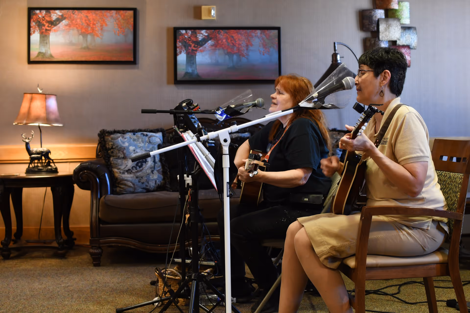 Two women seated and playing guitars while singing into microphones in a cozy living room setting with a brown leather couch, decorative pillows, a side table with a lamp and deer figurine, and framed pictures of autumn trees on the wall.