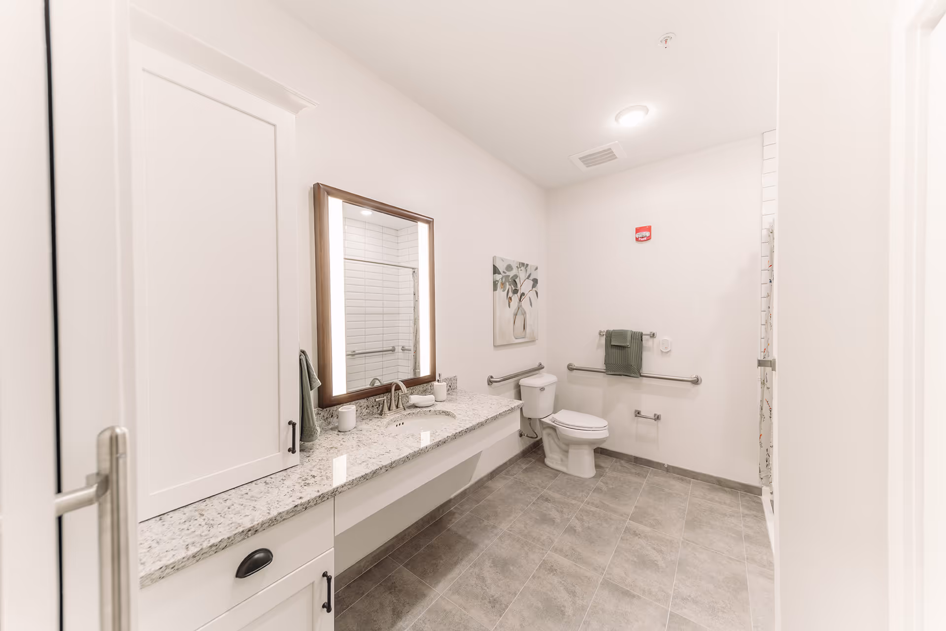 A spacious, modern bathroom with light-colored walls and gray tiled floor. It features a long granite countertop with a sink, a large illuminated mirror, a toilet with grab bars on the walls, a towel rack with a green towel, and a shower area with a curtain. There is also a decorative painting on the wall.