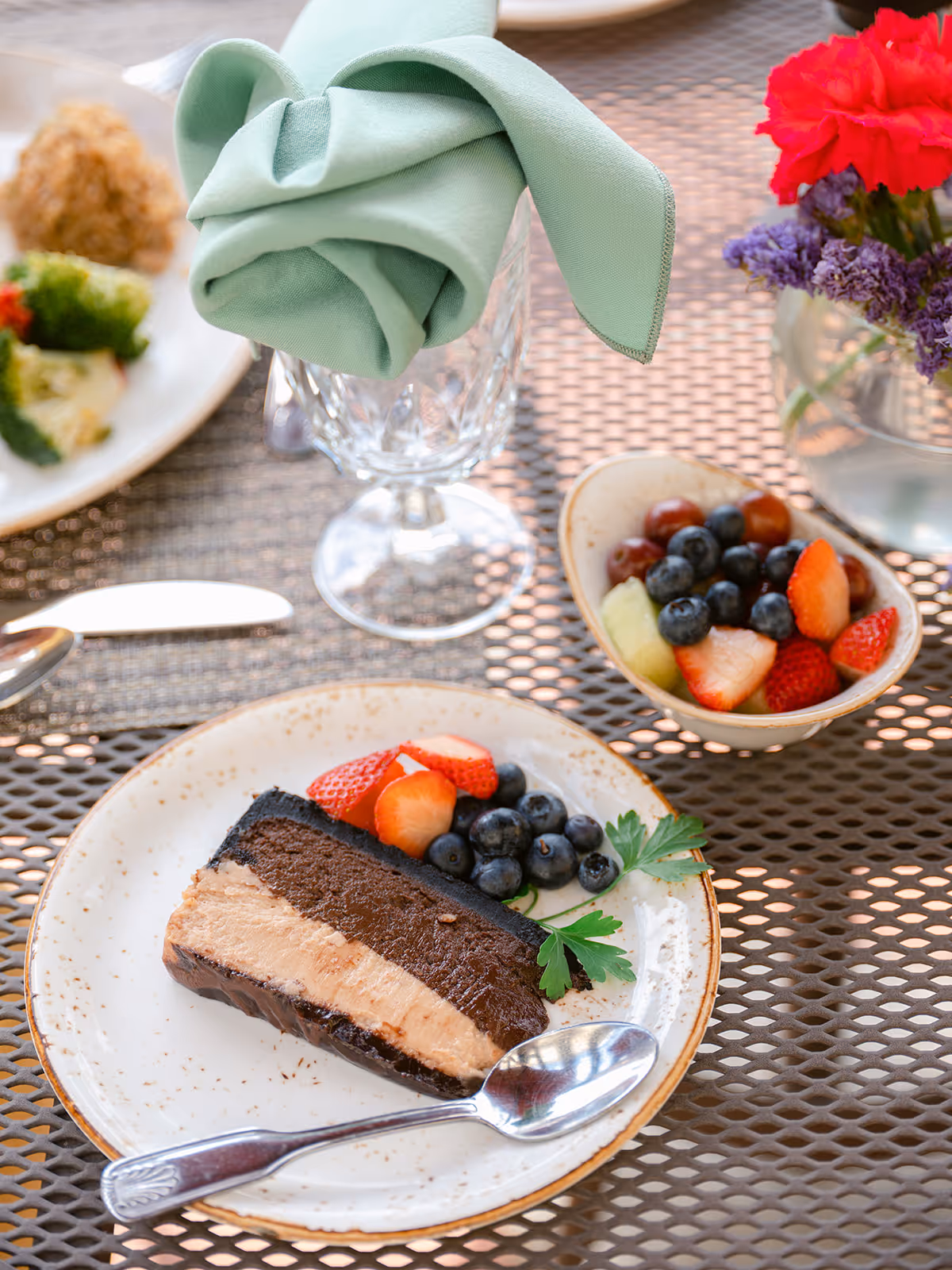 A plated dessert with a slice of layered chocolate mousse cake garnished with fresh strawberries and blueberries, accompanied by a small bowl of mixed fruit including grapes, blueberries, strawberries, and melon. A green cloth napkin is elegantly folded in a glass, and a small vase with red and purple flowers is visible on the table.