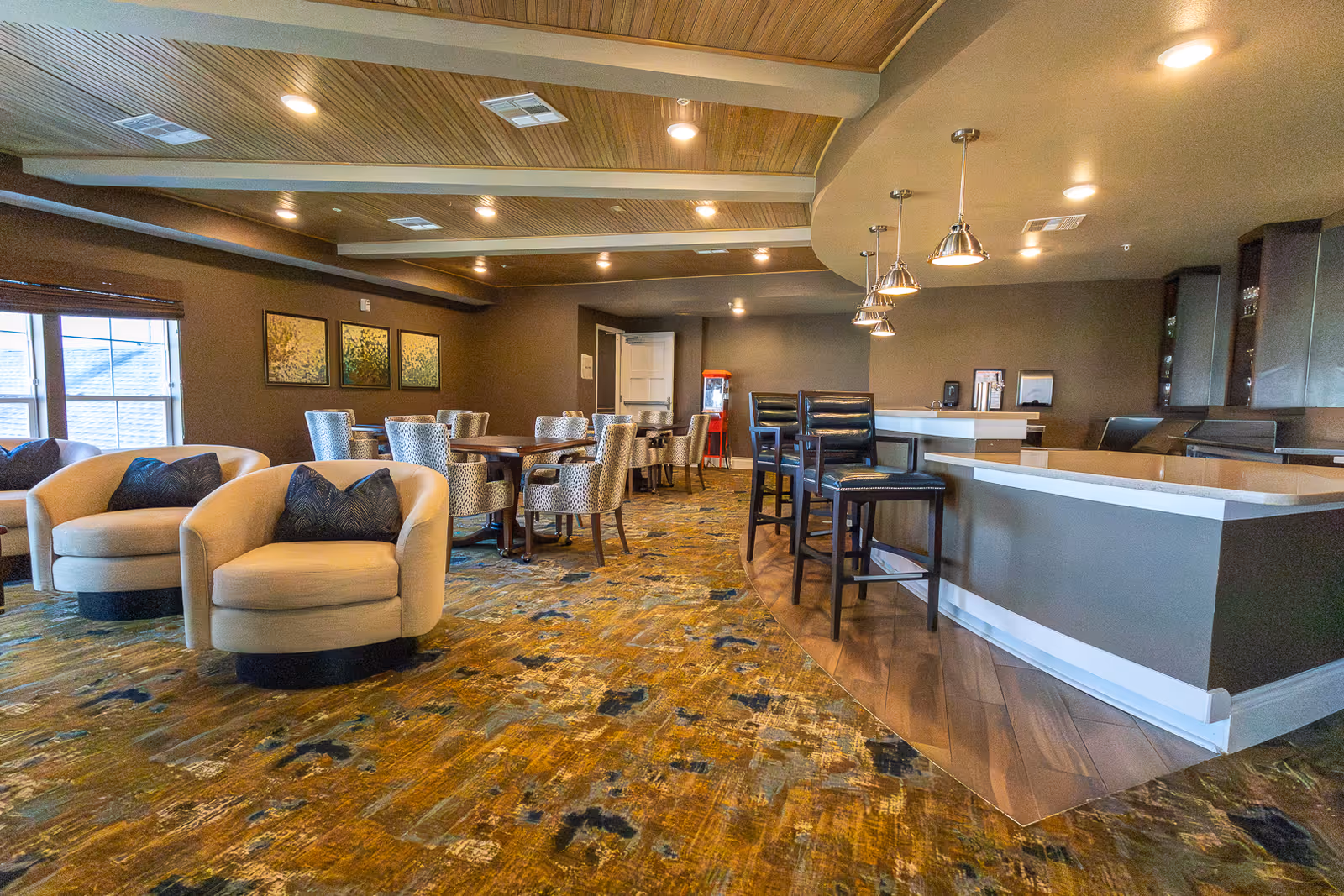A spacious senior living common area with a seating arrangement including beige armchairs with dark cushions, several dining tables with patterned chairs, and a kitchen counter with high stools and pendant lights overhead. The room has a patterned carpet, wooden ceiling beams, and wall art near the windows.