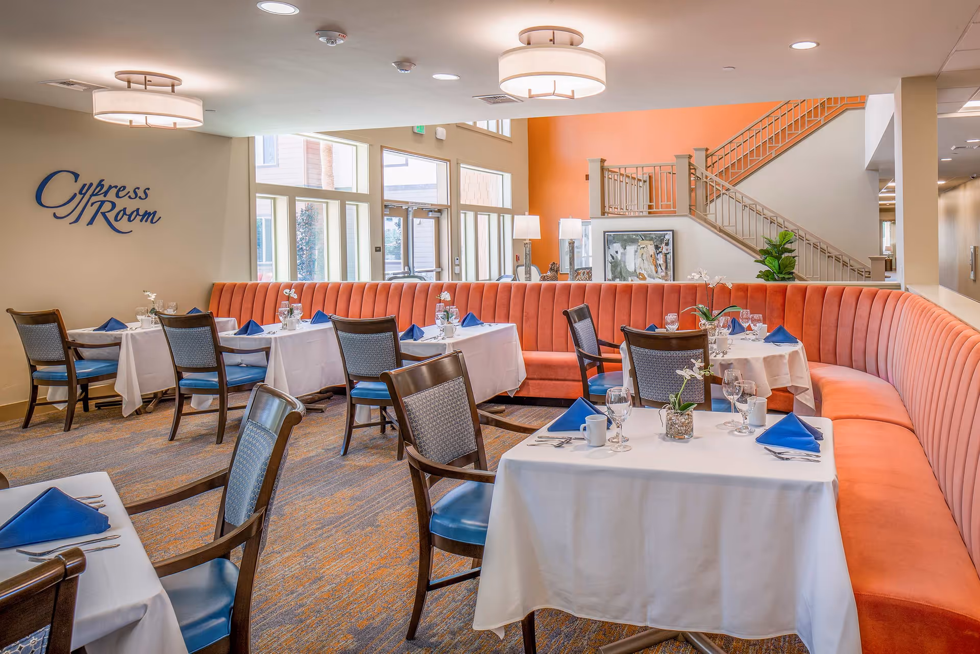 Dining room named Cypress Room with tables set for meals, featuring white tablecloths, blue napkins, glassware, and small flower centerpieces. The room has an orange curved banquette seating along the wall, wooden chairs with blue cushions, large windows letting in natural light, and a staircase in the background.