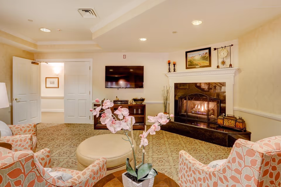 A cozy living room with patterned armchairs arranged around a round ottoman and a small table with a pink orchid plant. A fireplace with a fire burning is on the right wall, topped with decorative candles, a clock, and a framed landscape painting. A flat-screen TV is mounted on the wall above a wooden cabinet. Double doors are open to an adjacent room with a framed picture on the wall.