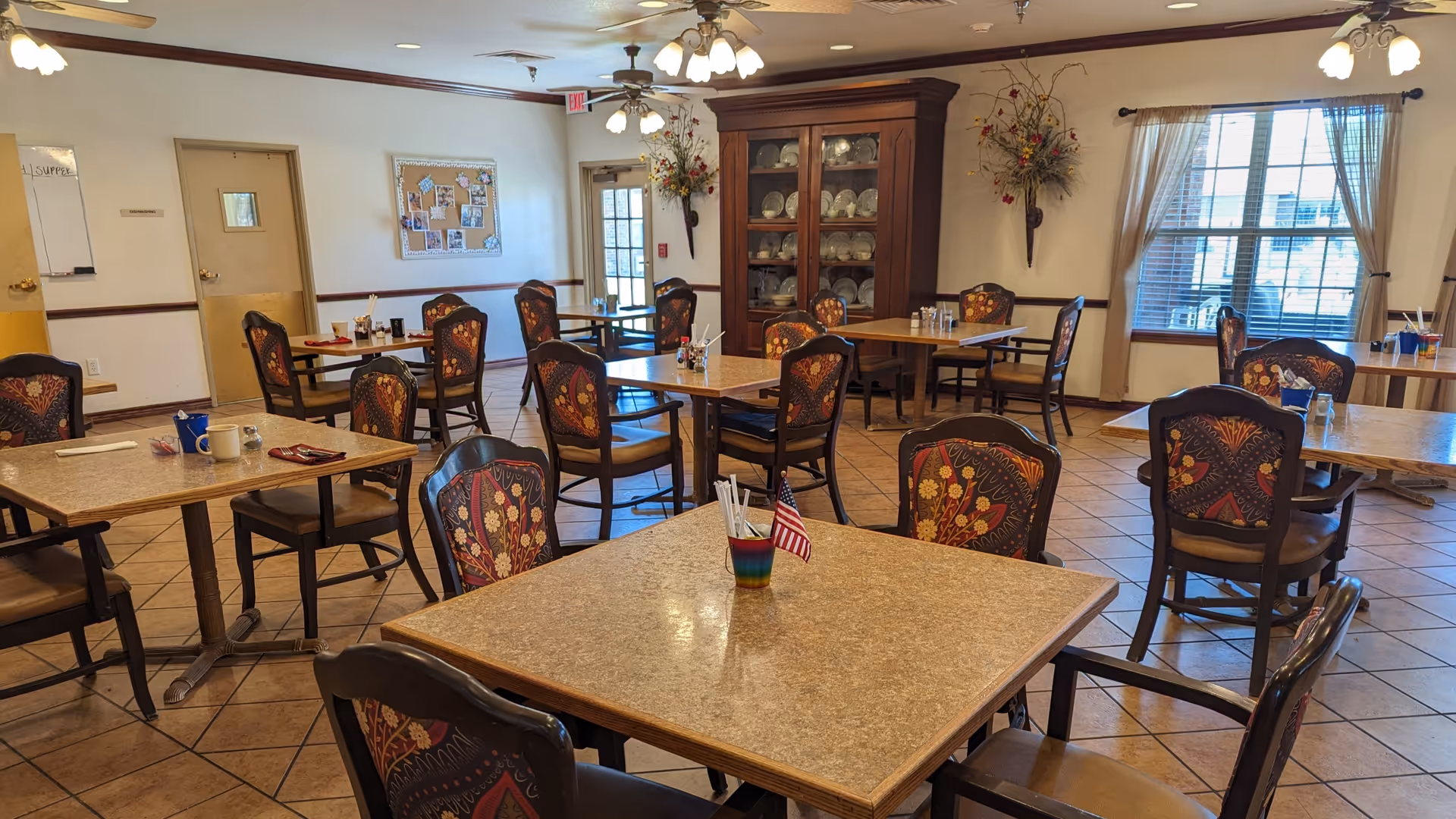 Communal dining room with multiple square tables and patterned chairs, ceiling fans, a china cabinet, and an American flag centerpiece.